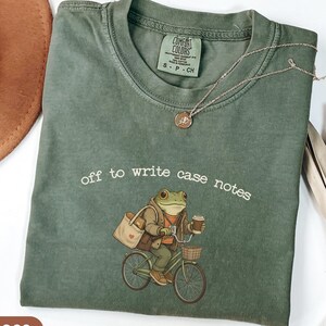 Könnte beinhalten: Moosgrünes Comfort Colors T-Shirt mit dem Schriftzug "off to write case notes" über einer Froschfigur auf einem Fahrrad. Der Frosch trägt eine Jacke und hält eine Kaffeetasse. Eine silberne Halskette mit Anhänger befindet sich auf dem Shirt.