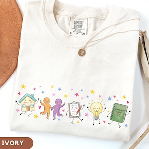 May include: An ivory short-sleeve t-shirt featuring a graphic print of a smiling house, orange and purple figures, a clipboard with a pencil, a lightbulb, and a green book labelled Community Guide, all surrounded by small colourful stars and hearts.