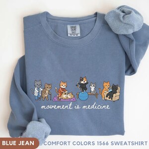 May include: A blue jean colored crewneck sweatshirt featuring a graphic of various cats engaged in physical therapy and exercise activities. Below the cats, the text reads movement is medicine. The garment has a Comfort Colors brand label inside.