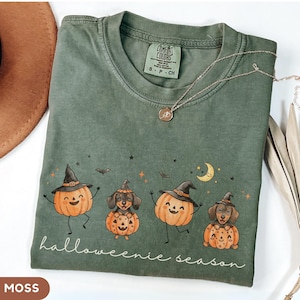 May include: A moss green short-sleeve t-shirt featuring a graphic of two smiling jack-o'-lanterns and two dachshunds wearing witch hats. The text "halloweenie season" is printed below the graphic. The shirt has a Comfort Colors brand label.