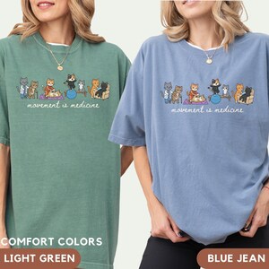 May include: A light green and a blue jean t-shirt, each featuring a graphic illustration of various cats performing physical therapy exercises with the text "movement is medicine" printed in white cursive script below the cat characters.