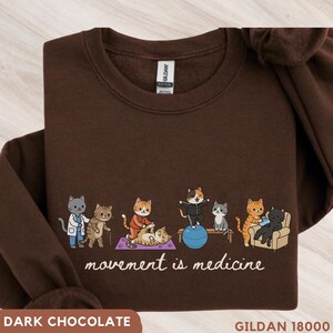 May include: A dark brown crewneck sweatshirt featuring a graphic of several cartoon cats exercising with the text "movement is medicine" printed below. The cats are shown performing various physical activities including stretching and using a blue ball.