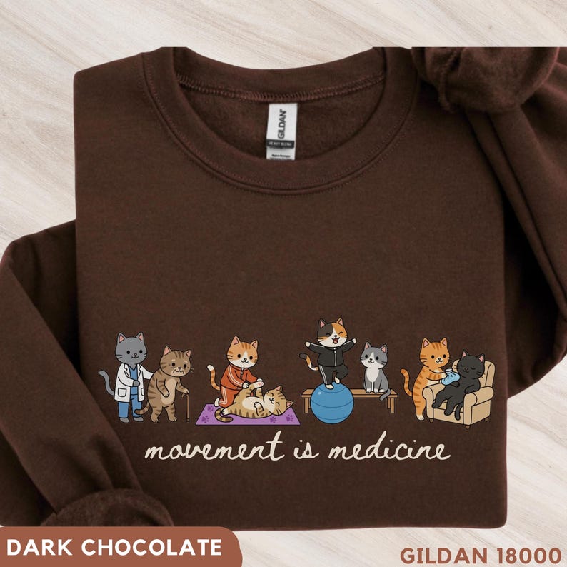 May include: A dark brown crewneck sweatshirt featuring a graphic of several cartoon cats exercising with the text "movement is medicine" printed below. The cats are shown performing various physical activities including stretching and using a blue ball.