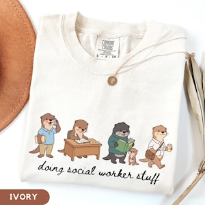 May include: An ivory t-shirt featuring a graphic of four cartoon otters performing various tasks, with the text "doing social worker stuff" printed below. The shirt has a Comfort Colors brand label at the neck and a crew neckline design.