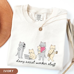May include: An ivory t-shirt featuring a graphic of four cats engaged in various office tasks with the text "doing social worker stuff" printed below. The shirt has a Comfort Colors brand label at the neck and a thin gold-toned chain necklace.