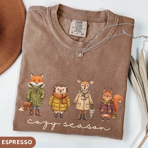 May include: A brown crewneck t-shirt featuring a graphic of a fox, hedgehog, deer, and squirrel wearing winter clothing. The text "cozy season" is printed below the animals in white script. The shirt has a Comfort Colors brand label at the neck.
