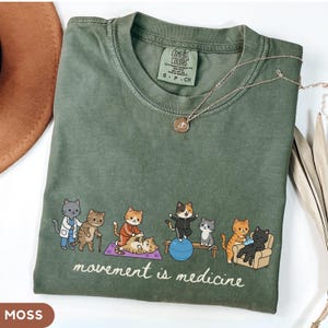 May include: An olive green t-shirt featuring a graphic of cartoon cats exercising and receiving medical care. The text "movement is medicine" is printed below the cats. The shirt has a Comfort Colors label indicating a size small.