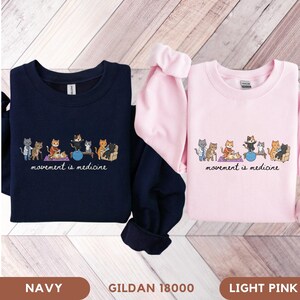 May include: A navy blue sweatshirt and a light pink sweatshirt, each featuring a graphic of cartoon cats performing various physical therapy exercises with the printed text movement is medicine below the illustration on the front of the garments.