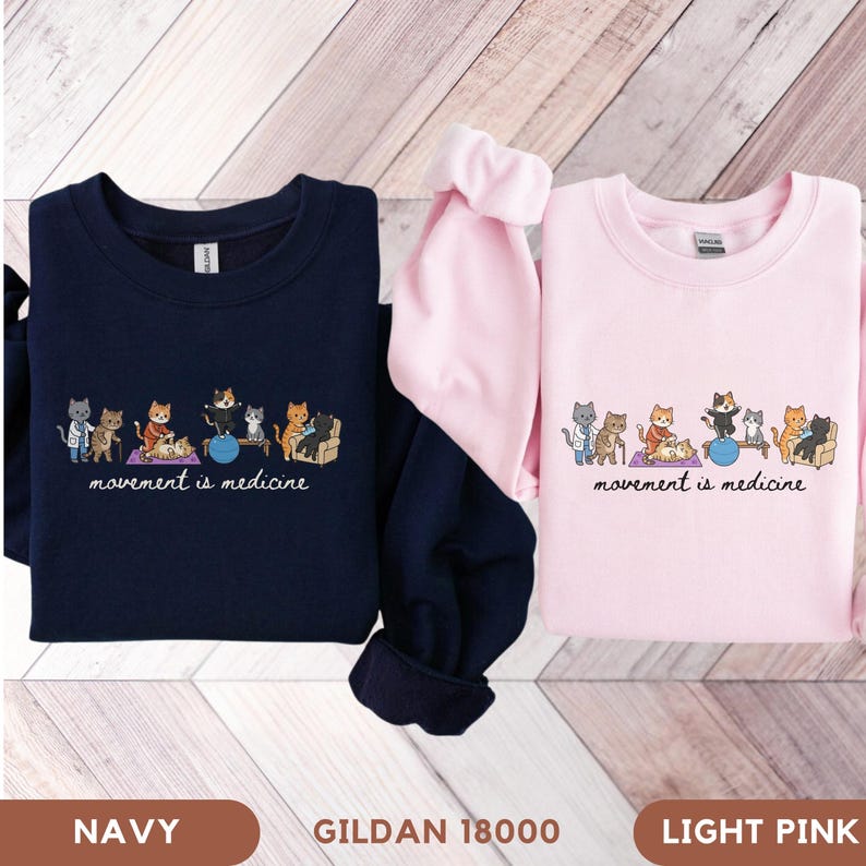 May include: A navy blue sweatshirt and a light pink sweatshirt, each featuring a graphic of cartoon cats performing various physical therapy exercises with the printed text movement is medicine below the illustration on the front of the garments.