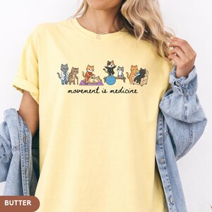 May include: A pale yellow t-shirt featuring a graphic illustration of various cats performing physical therapy exercises, including stretching and using a balance ball. Below the cats, the text reads, movement is medicine, in black script.