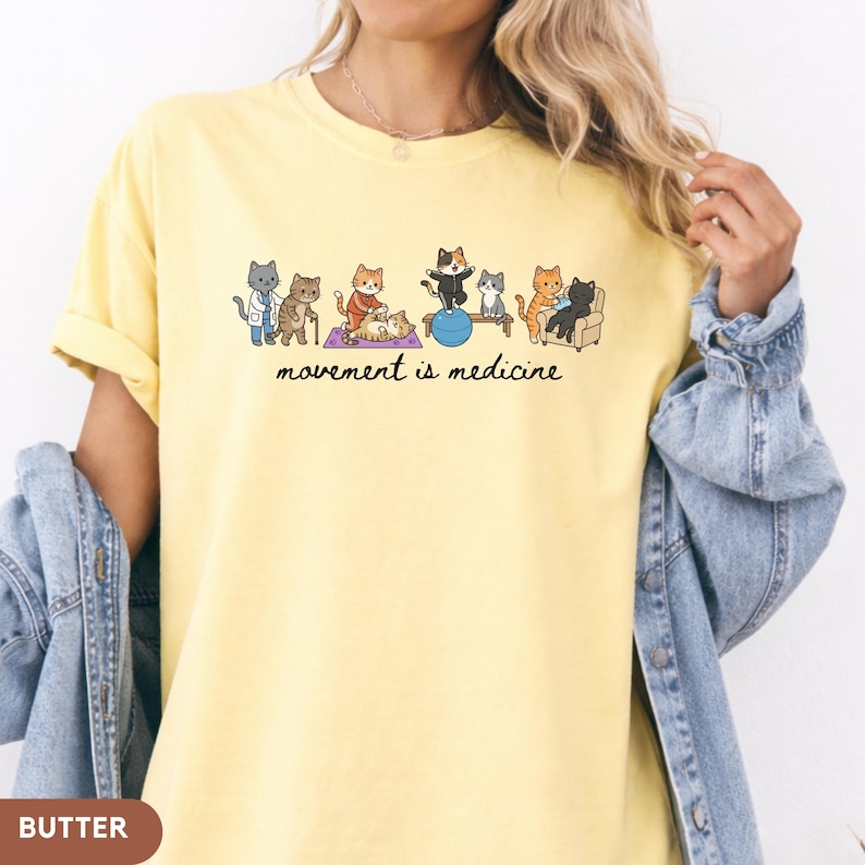 May include: A pale yellow t-shirt featuring a graphic illustration of various cats performing physical therapy exercises, including stretching and using a balance ball. Below the cats, the text reads, movement is medicine, in black script.