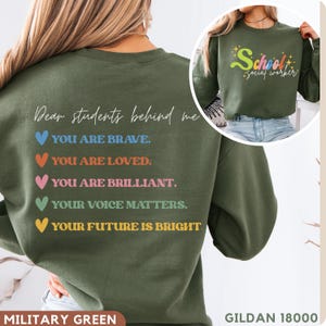 Könnte beinhalten: Militärgrünes Sweatshirt mit dem Text "Dear students behind me" und inspirierenden Botschaften. Auf der Rückseite des Sweatshirts stehen die Sätze "YOU ARE BRAVE," "YOU ARE LOVED," "YOU ARE BRILLIANT," "YOUR VOICE MATTERS" und "YOUR FUTURE IS BRIGHT." Auf der Vorderseite steht der Text "School social worker."