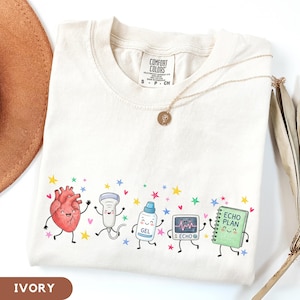 Puede incluir: Una camiseta de color marfil con un estampado gráfico de cinco personajes de dibujos animados: un corazón rojo, una sonda de ultrasonido, un bote de gel, un monitor de eco y un cuaderno verde etiquetado ECHO PLAN, rodeados de estrellas y corazones.