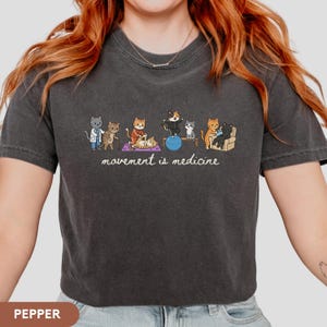 May include: A charcoal gray t-shirt featuring a graphic of several cats performing physical therapy exercises. The illustration includes cats on a yoga mat, a balance ball, and a chair. Below the cats, the text reads movement is medicine.