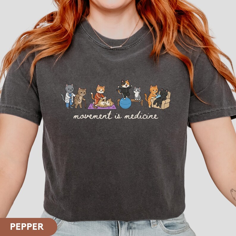 May include: A charcoal gray t-shirt featuring a graphic of several cats performing physical therapy exercises. The illustration includes cats on a yoga mat, a balance ball, and a chair. Below the cats, the text reads movement is medicine.