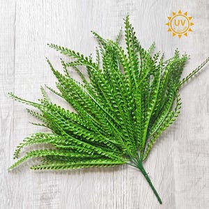 May include: Bright green artificial fern plant with small, closely spaced leaves. The plant has a natural appearance and is suitable for indoor or outdoor use. The image also includes a UV protection symbol.