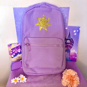 May include: A lavender backpack with a gold glitter sun design on the front. The backpack has a front pocket with a zipper and adjustable straps. The background includes purple and white star-patterned paper and a drawing of a tower.