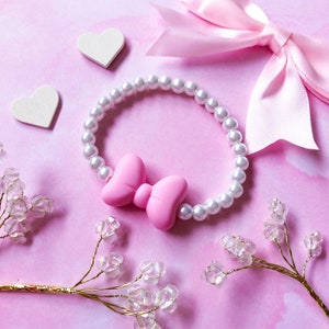 May include: A pink bow charm attached to a white pearl bracelet. The bracelet is on a pink background with two white hearts and two sprigs of gold and crystal embellishments.