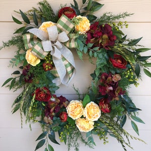 May include: A green and white checkered ribbon bow with a burlap center decorates a wreath made of greenery, burgundy and yellow flowers.