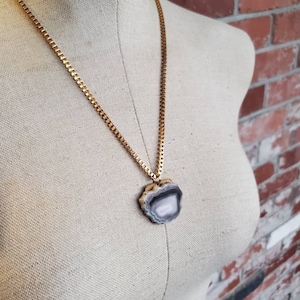 May include: A gold-toned chain necklace with a pendant featuring a polished, gray and white agate stone. The stone is irregularly shaped with a natural, layered pattern. The necklace is displayed on a mannequin against a brick wall.