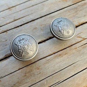 May include: Two silver-toned buttons with a raised design featuring a crown, crossed swords, and a shield. The buttons have a textured surface and a beaded edge.