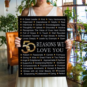 May include: A black and gold sign with the text "50 REASONS WE LOVE YOU." The sign lists qualities such as "Kind," "Passionate," and "Honest & Beautiful." The sign is held by a person.