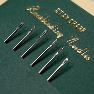 Puede incluir: Un sobre de cartulina verde con detalles en pan de oro y el texto "STAY SHARP Bookbinding Needles" en letras doradas. El sobre contiene un juego de agujas de encuadernación plateadas.