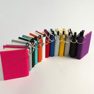 Puede incluir: Una colección de libros en miniatura dispuestos en semicírculo, cada uno con una cubierta de tela de color sólido en tonos rosa, gris, turquesa, naranja, blanco, rojo, azul, amarillo y morado, con una anilla de metal plateado en el lomo.