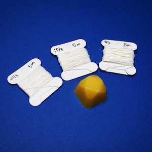 Pode incluir: Três rolos de fio branco, cada um etiquetado com uma fração e "5m". Um pequeno bloco hexagonal de cera de abelha amarelo fica no centro.