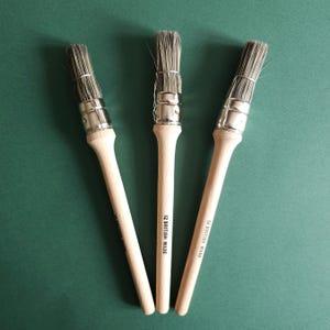 Może przedstawiać: Trzy pędzle z jasnymi drewnianymi trzonkami i srebrnymi metalowymi skuwkami. Włosie jest szare i trzymane razem drutem. Na trzonku nadrukowano napis "12 BRITISH MADE". Pędzle ułożone są na zielonej powierzchni.