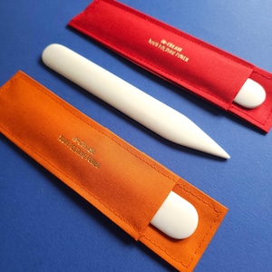 Peut inclure: Deux limes à ongles blanches dans des étuis en velours orange et rouge. Les étuis portent l'inscription dorée "Increase Your Folding Power".