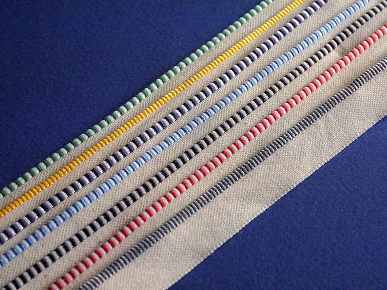 May include: A close-up of a white fabric with different colored stripes sewn along the edge. The stripes are green, yellow, navy blue, light blue, black, red, gray, and dark blue.