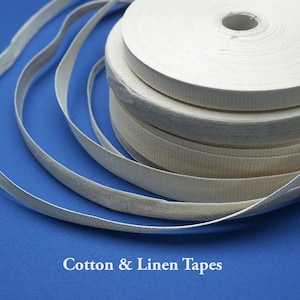 Puede incluir: Primer plano de varios rollos de cintas de algodón y lino blanquecinas. Las cintas están dispuestas sobre una superficie azul. El texto "Cotton & Linen Tapes" está en la parte inferior de la imagen.