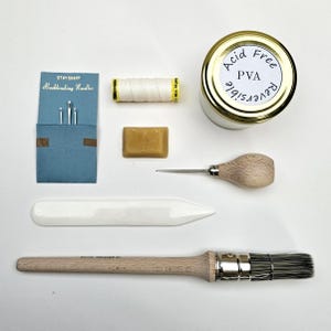 Op de afbeelding: Een verzameling boekbindgereedschap, waaronder naalden, draad, een klein blokje was, een vouwbeen, een borstel en een potje met het label "Acid Free PVA Reversible". De gereedschappen zijn op een wit oppervlak geplaatst.