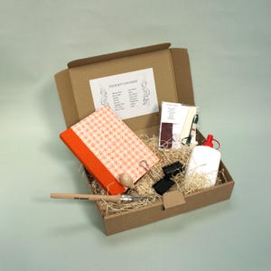 May include: An open cardboard box filled with craft supplies, including a patterned notebook, glue, and a wooden tool. The box contains a label that reads "YOUR KIT CONTAINS". The notebook has an orange spine and a red and white floral pattern.
