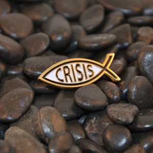 May include: A gold-colored fish-shaped pin with a white background and the word "CRISIS" written in black letters.