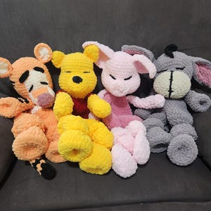 May include: Four handmade crochet plush toys of cartoon characters, including a yellow bear, an orange tiger, a pink bunny, and a gray donkey. The toys are soft and cuddly, with closed eyes, suggesting they are sleeping. The toys are resting on a gray surface.