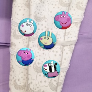 May include: Five colorful fabric buttons with cartoon characters from the children's show Peppa Pig. The buttons are attached to white ribbon and are on a white and purple fabric background with a star and crescent moon pattern.
