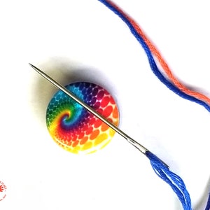 May include: A rainbow swirl design needle minder with a silver needle threaded through it. Two strands of embroidery floss, one blue and one orange, are attached to the needle.