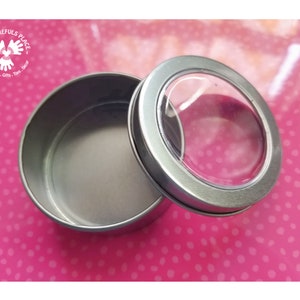 May include: A silver round tin container with a clear plastic lid. The lid has a round window to see inside the container.