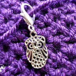 Może przedstawiać: Srebrny znacznik ściegu w kształcie sowy z srebrnym klipsem. Znacznik znajduje się na fioletowym materiale dzianym na szydełku.