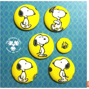 Könnte beinhalten: Satz mit sechs runden gelben Stoffknöpfen mit einem Cartoon-Bild von Snoopy, dem Beagle aus dem Comic Peanuts. Die Knöpfe sind mit schwarzen und weißen Umrissen verziert und haben einen weißen Hintergrund. Die Knöpfe sind in einem Kreis auf einem blauen Hintergrund mit einem weißen Rautenmuster angeordnet.