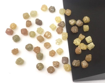 May include: A selection of raw, unpolished diamonds in shades of yellow, brown, and green. The diamonds are spread out on a white background, with some near a black, angled container. The gemstones have a natural, irregular form.