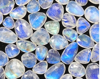 Puede incluir: Una colección de piedras preciosas de piedra lunar facetadas. Las piedras varían en forma y tamaño, mostrando una gama de colores que van del transparente al blanco lechoso, con destellos de azul y tonos iridiscentes. Las piedras están dispuestas sobre una superficie negra.