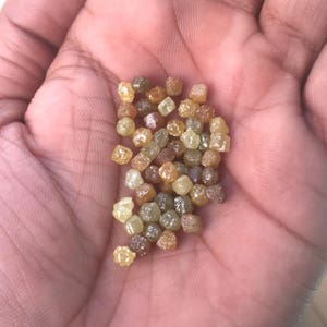 Puede incluir: Un pu&ntilde;ado de diamantes en bruto, sin cortar, en varios tonos de amarillo, marr&oacute;n y transparente. Las peque&ntilde;as piedras en forma de cubo se sostienen en la palma de la mano, mostrando su estado natural y sin pulir. Los diamantes tienen un aspecto ligeramente texturizado.