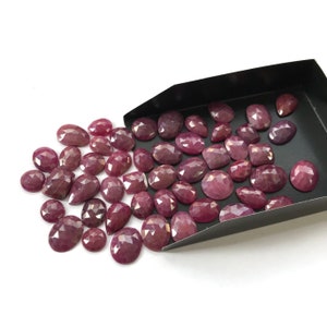 Könnte beinhalten: Eine Sammlung facettierter, tiefroter Edelsteine in verschiedenen Formen, darunter oval, birnenförmig und kissenförmig. Die Edelsteine sind auf einer weißen Oberfläche verstreut, einige fallen aus einer schwarzen Schale. Die Steine haben eine polierte, reflektierende Oberfläche.