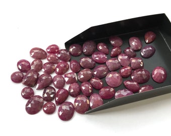 Puede incluir: Una colección de gemas facetadas de color rojo intenso en varias formas, incluyendo óvalos, peras y cojines. Las gemas están dispersas sobre una superficie blanca, algunas se derraman de una bandeja negra. Las piedras tienen una superficie pulida y reflectante.