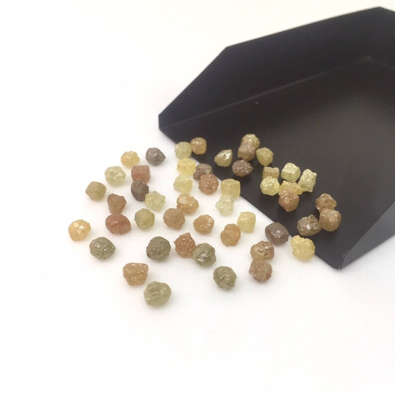 Puede incluir: Una colecci&oacute;n de diamantes en bruto, sin cortar, en varios colores, incluyendo amarillo, marr&oacute;n y verde, dispersos sobre una superficie blanca y una bandeja negra. Las piedras preciosas tienen una forma natural e irregular.