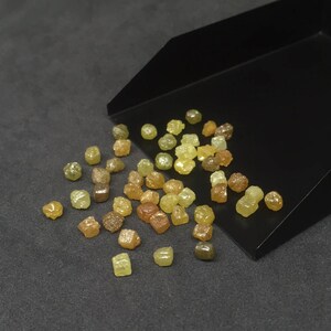 Puede incluir: Una colecci&oacute;n de diamantes en bruto sin cortar en varios tonos de amarillo, marr&oacute;n y verde. Las piedras preciosas est&aacute;n esparcidas sobre una superficie gris oscuro, con algunas saliendo de una bandeja negra. Los diamantes tienen una forma natural e irregular.