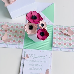 May include: A white box with a pink bow on top. The box is open and inside there is a pop-up card with a quote in Italian that says "Mamma la parola più bella sulle labbra dell'umanità. Khalil Gibran". The card has pink and red paper flowers and butterflies.